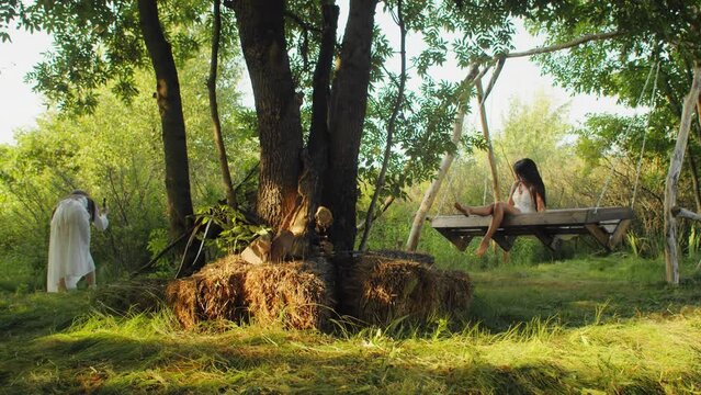 Person in a white robe photographing a woman relaxing on an oversized swing in a whimsical forest setting. Creative storytelling and fantasy concept. Design for film and photography project promotions