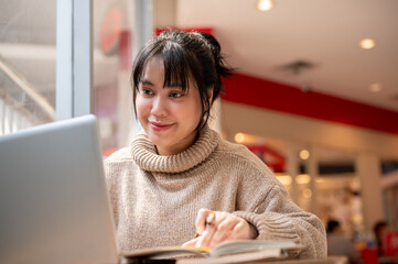 An attractive Asian woman in a comfy sweater is working remote at a cafe, working on her laptop.
