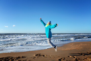 A young girl jumps on the seashore. Happy blonde woman in warm clothes in the winter season on a sunny day. Freedom and activity.