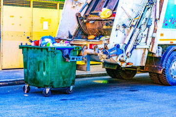 A trash can with waste in selective focus stands in front of a garbage truck before loading and...