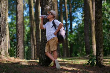 A man leans against a tree trunk while drinking water from a plastic bottle; a backpack on his shoulder