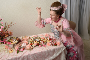 Close up portrait of cute female model wearing an opulent pink gown, the costume of a historical French baroque nobility.  sitting table with sweet cakes and indulgent food feast.
 - obrazy, fototapety, plakaty