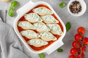Raw conchiglioni pasta stuffed ricotta cheese and spinach with tomato sauce in a white baking dish on a gray concrete background.
