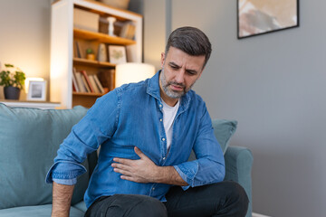 Fototapeta premium Terrible stomachache. Frustrated handsome young man hugging his belly and keeping eyes closed while lying on the couch at home