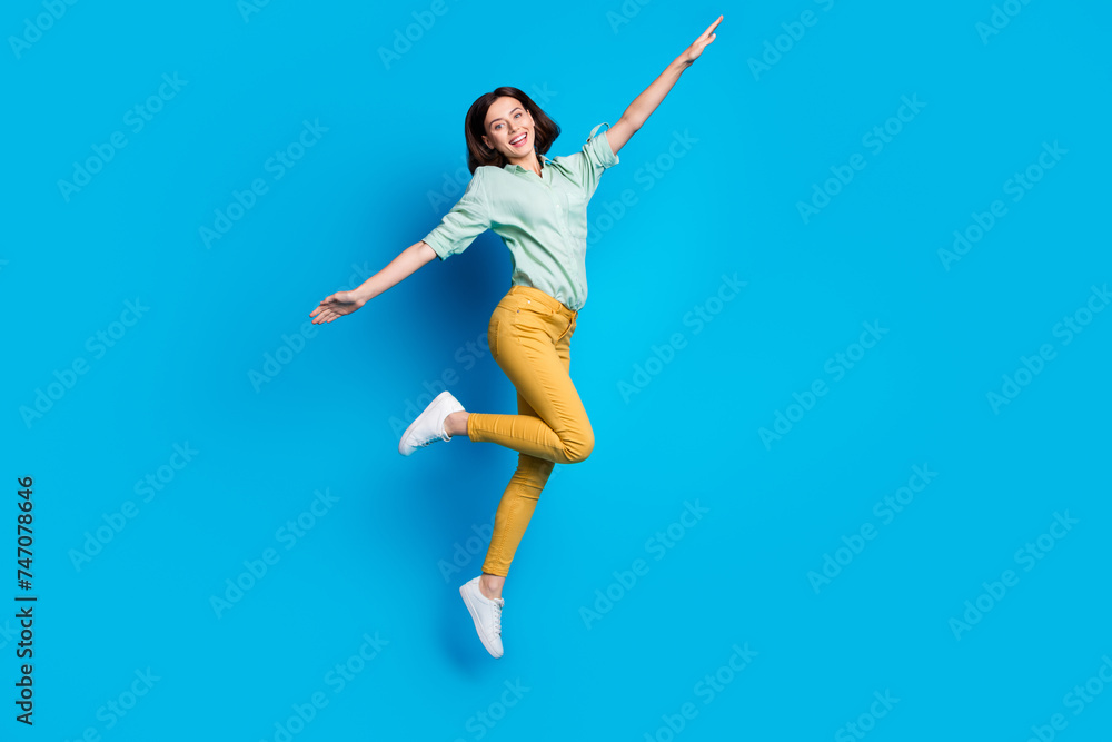 Poster Full length photo of girlish woman dressed teal shirt yellow trousers jumping hold arms like wings isolated on blue color background