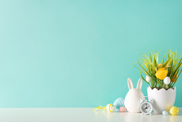 Festive Easter display, featuring side view of table with figurine bunny, assorted colored eggs, shell shaped flowerpot with grass and flowers, alarm clock, against a pastel blue wall, space for copy