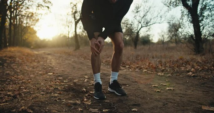 A guy an athlete in a black sports uniform while jogging in the forest holds his knee and rubs it because he feels damage and injury