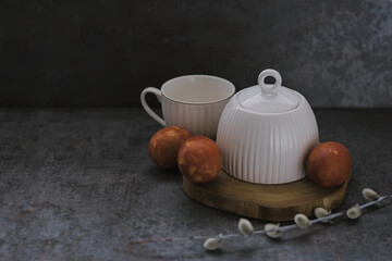 Festive Easter spring composition with white ceramic tea pair and red eggs on wooden podium with willow branch and handmade paper rabbit on dark background