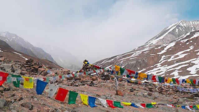 Buddhist prayer flags tying on rocks on mountains at Baralacha La Pass in Himachal Pradesh, India. Sacred religious belief of god. Beautiful Himalaya mountains with snow and moody clouds in sky. 