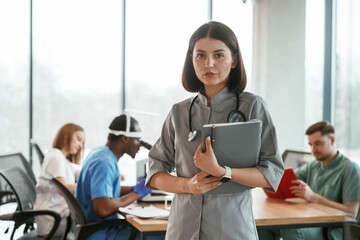 Woman in grey uniform is standing with notepad. Group of doctors are together indoors