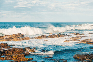 Sea waves crashing on rocks in sunny day against cloudy sky