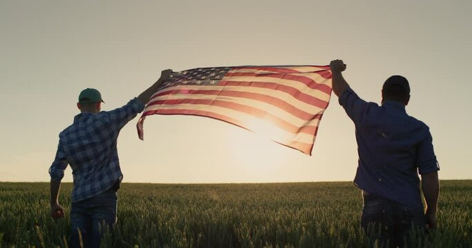 Two men raise up the US flag against the backdrop of a wheat field at sunset