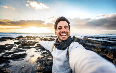 Handsome young man taking selfie pic with smart mobile phone outdoors - Traveler guy smiling at camera with sunset on background - Traveling life style and technology concept - Powered by Adobe