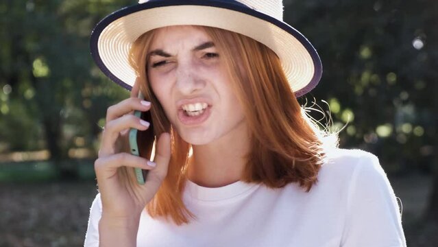 Angry pissed off teenage girl with red hair wearing straw hat and pink earphones talking fiercely on mobile phone
