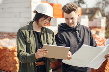 Stylish woman architect with tablet  and contractor man checking blueprints at construction site. Young engineer or construction workers in hardhat looking at plans of new modern house