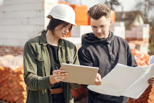 Stylish woman architect with tablet and contractor man checking blueprints at construction site. Young engineer or construction workers in hardhat looking at plans of new modern house - Powered by Adobe
