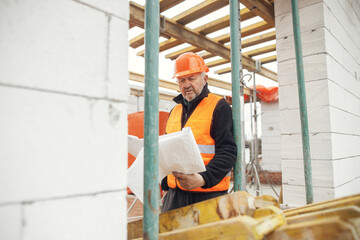 Senior man engineer or construction worker in hardhat looking at blueprints at building new modern house. Male architect or contractor checking plans at construction site. Copy space