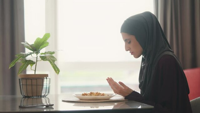 Muslim woman in headscarf opening palms and praying before dinner, traditions