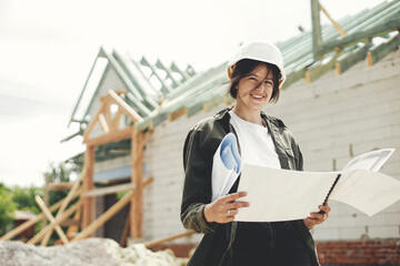 Stylish happy woman architect checking blueprints against modern farmhouse construction site. Young female engineer or construction worker in hardhat with plans of new modern house