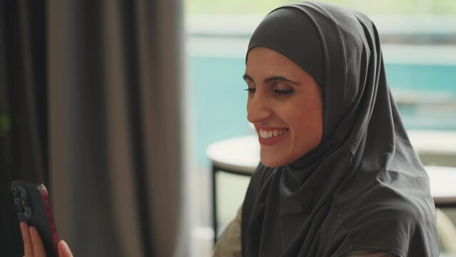Positive young Arab woman in headscarf having online video call on smartphone