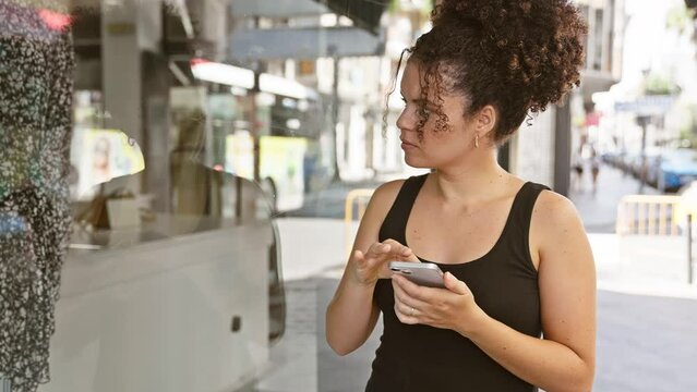 Young hispanic woman with curly hair using smartphone on urban street, casual, technology, outdoors.