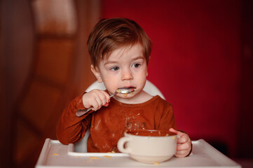 A young child focuses intently on scooping the remnants of a meal from a bowl, oblivious to the scattered food on the highchair tray and their own face. 