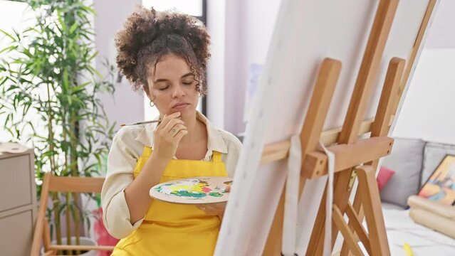 Thoughtful hispanic artist with curly hair, sitting in art studio, hand on chin, pensive expression on her beautiful face, questioning and doubt evident, happily deliberating a creative solution.