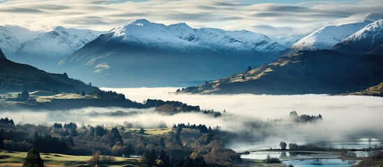 The image captures a breathtaking view of a valley with snow-capped mountains in the background on a misty morning in Canterbury, New Zealand. The valley overlooks Robinsons Bay on Bank Peninsula. - obrazy, fototapety, plakaty
