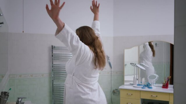 Slow motion. A woman enters the bathroom wearing a white bathrobe and pandiculation. A woman, yawning, approaches the wall mirror, rubs her eyes with her hands and looks into the mirror smiling