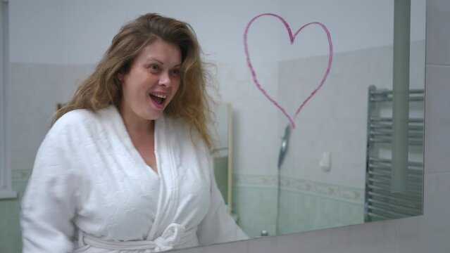Slow motion. A woman in a white bathrobe smiles and poses looking at herself in the mirror while standing in the bathroom. A heart is drawn on the mirror with red lipstick