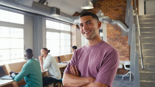 Portrait of smiling young businessman working in modern open plan office looking at camera and crossing arms with colleagues in background - shot in slow motion