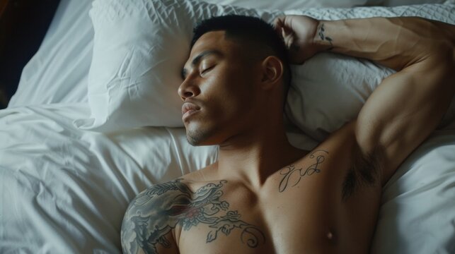 A tattooed man with short hair sleeping peacefully on his side with his arm resting on his head lying on a white bed with a pillow under his head.