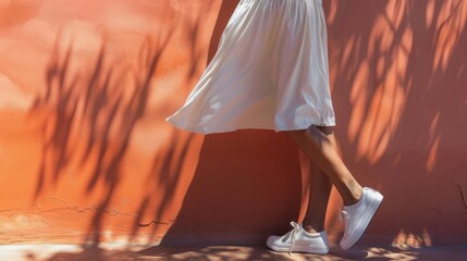A person wearing a white skirt and sneakers standing in front of an orange wall with shadows of leaves.