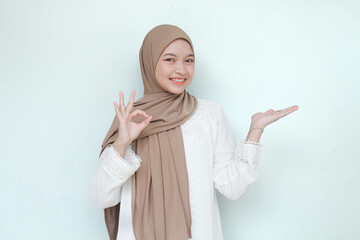 Young Asian Muslim woman wearing hijab, smiling and see camera while pointing and showing to copy space beside her with ok gesture. Isolated in gray background.