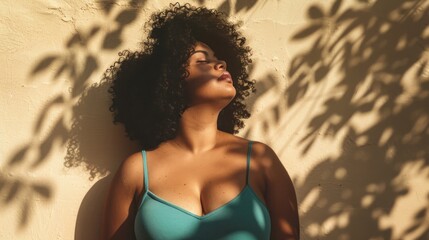 A woman with curly hair wearing a blue top leaning against a wall with her eyes closed basking in the sunlight filtering through leaves.