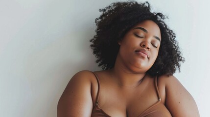 A woman with curly hair closed eyes and a relaxed expression wearing a brown top leaning against a white wall.