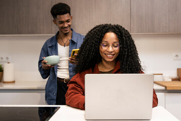 Diverse carefree couple planing together with computer and smartphone at home. Smiling man texting with phone while woman using laptop in the kitchen.