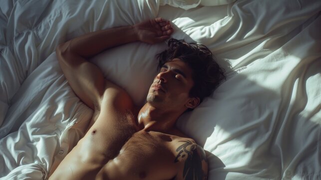 A shirtless man with tattoos lying on his side on a bed with his arm resting on his head in a relaxed and comfortable pose.