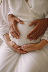 Fototapeta na wymiar Close up photo of pregnant woman in white dress and her husband holding their hands on pregnant belly. Love concept