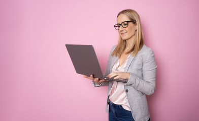 Charming clever lady in a beige formal smart casual shirt is standing on pink background with laptop in her hands