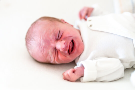 A close-up image capturing the raw emotion of a newborn baby crying, dressed in a soft white onesie against a gentle backdrop