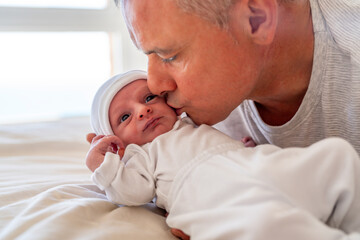 A father lovingly kisses his newborn's forehead while the baby looks on with wide eyes, creating a heartwarming family moment