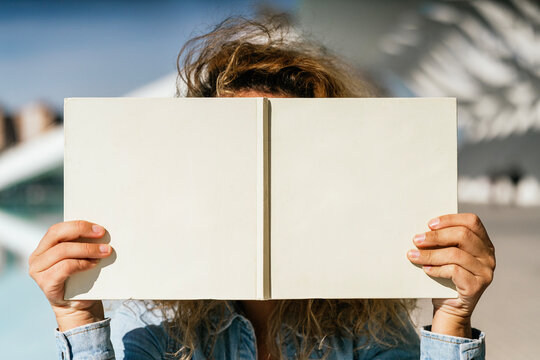 In a striking outdoor setting, anonymous person with hands that have been surgically adapted due to no thumbs holds an open book, covering their face