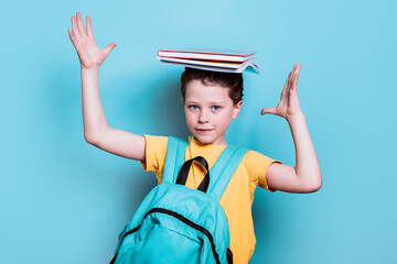 A school boy with a backpack strikes a playful pose, balancing a stack of books on his head against a light blue background