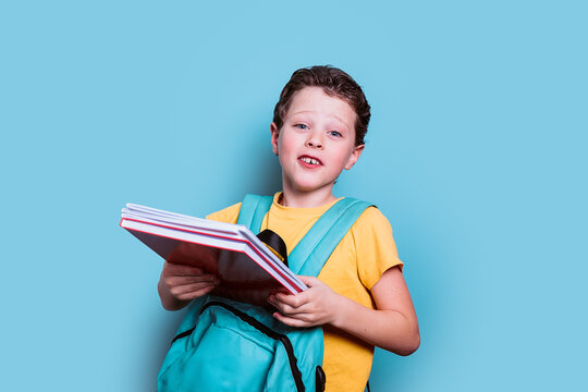 A young boy with a curious expression holds his school books while wearing a backpack, set against a refreshing blue background