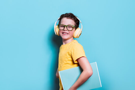 A happy young student in a yellow shirt and glasses wears headphones while holding a laptop, ready for a modern learning experience