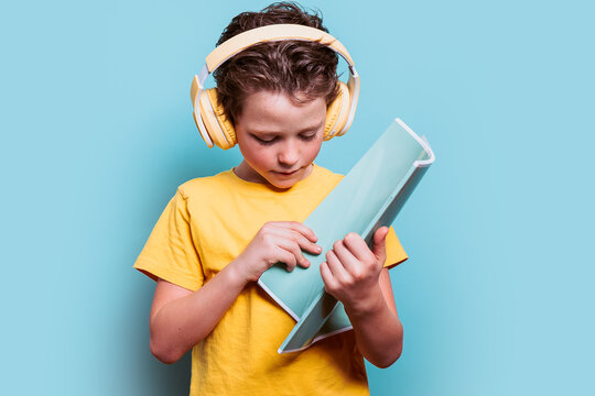 A young student deeply absorbed in reading a book while wearing large headphones, set against a cool blue background