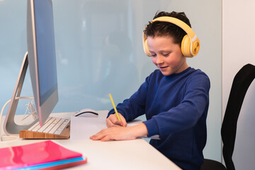 A dedicated child wearing headphones while writing notes at a desk with a computer, illustrating a modern multi-tasking study environment