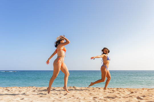 Two friends playfully dancing on the sand, sporting bright bikinis and hats, embodying the spirit of a carefree beach day
