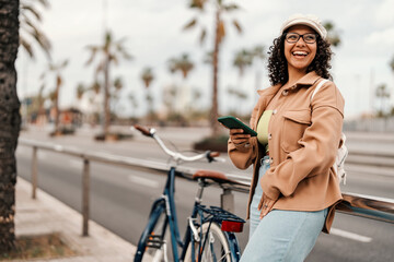 A happy gen z girl is leaning on the street with cellphone in hands near the bicycle.